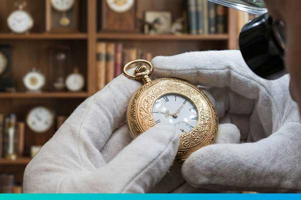 Nahaufnahme einer goldenen Taschenuhr die von einer Hand mit weißem Handschuh gehalten wird, ein Stück eines Gesichts mit einer lupe vor dem Auge ist auch zu sehen